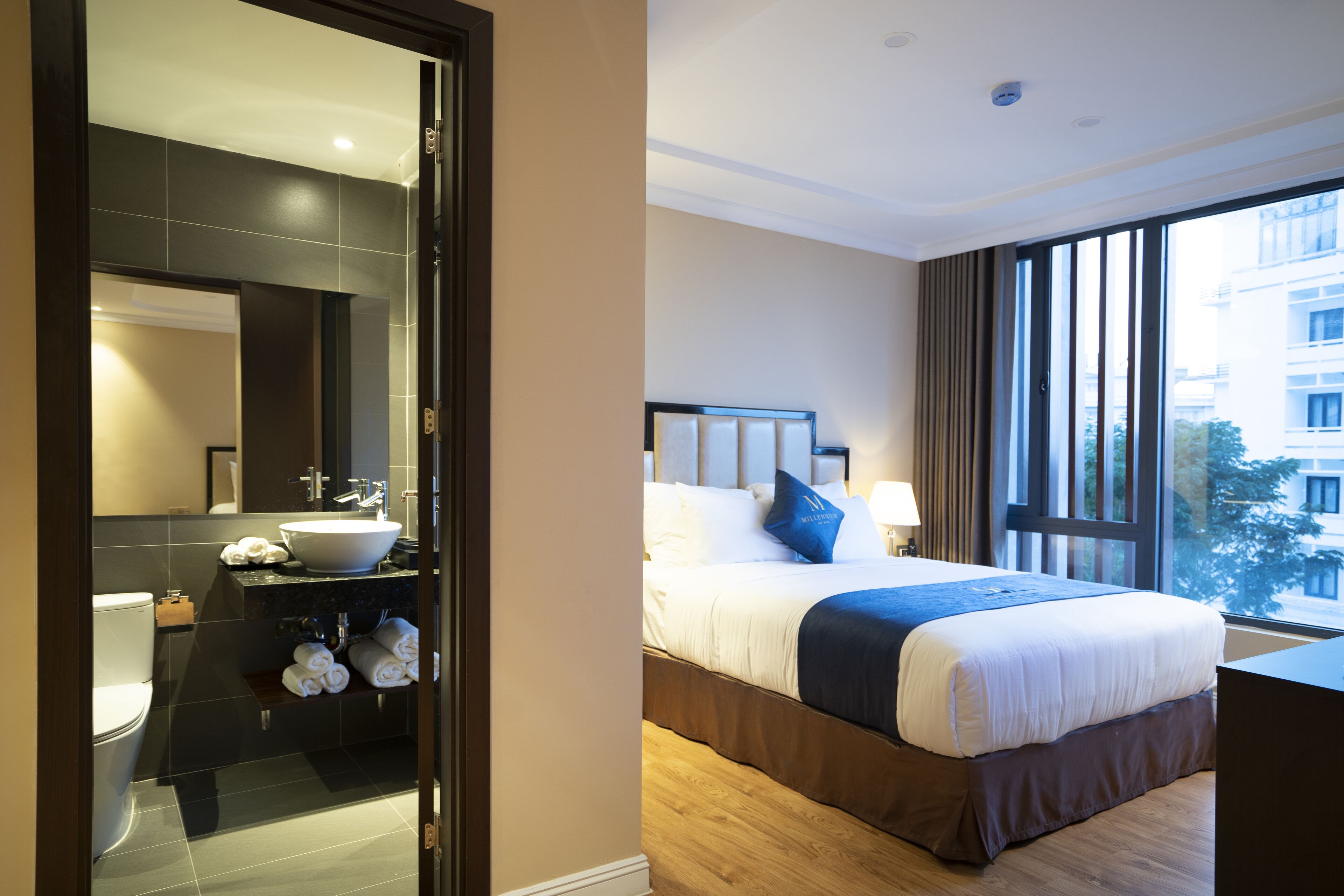 Una habitación de hotel moderna con una cama grande, un baño con lavabo y espejo, y vista a la ciudad por la ventana.