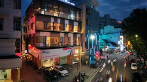 Un hotel moderno con un llamativo letrero 'Millennium', balcones, el logotipo visible de una agencia MB y una calle concurrida con coches y motocicletas.