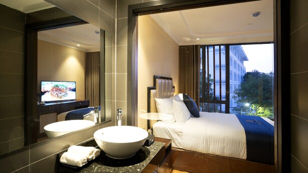 Una habitación de hotel moderna con una cama grande, un baño con lavabo y espejo, y vistas de la ciudad a través de amplios ventanales.