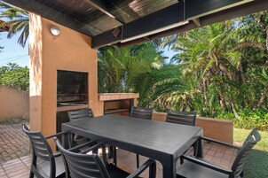 An outdoor patio with a table and chairs, a built-in grill, and lush tropical vegetation.