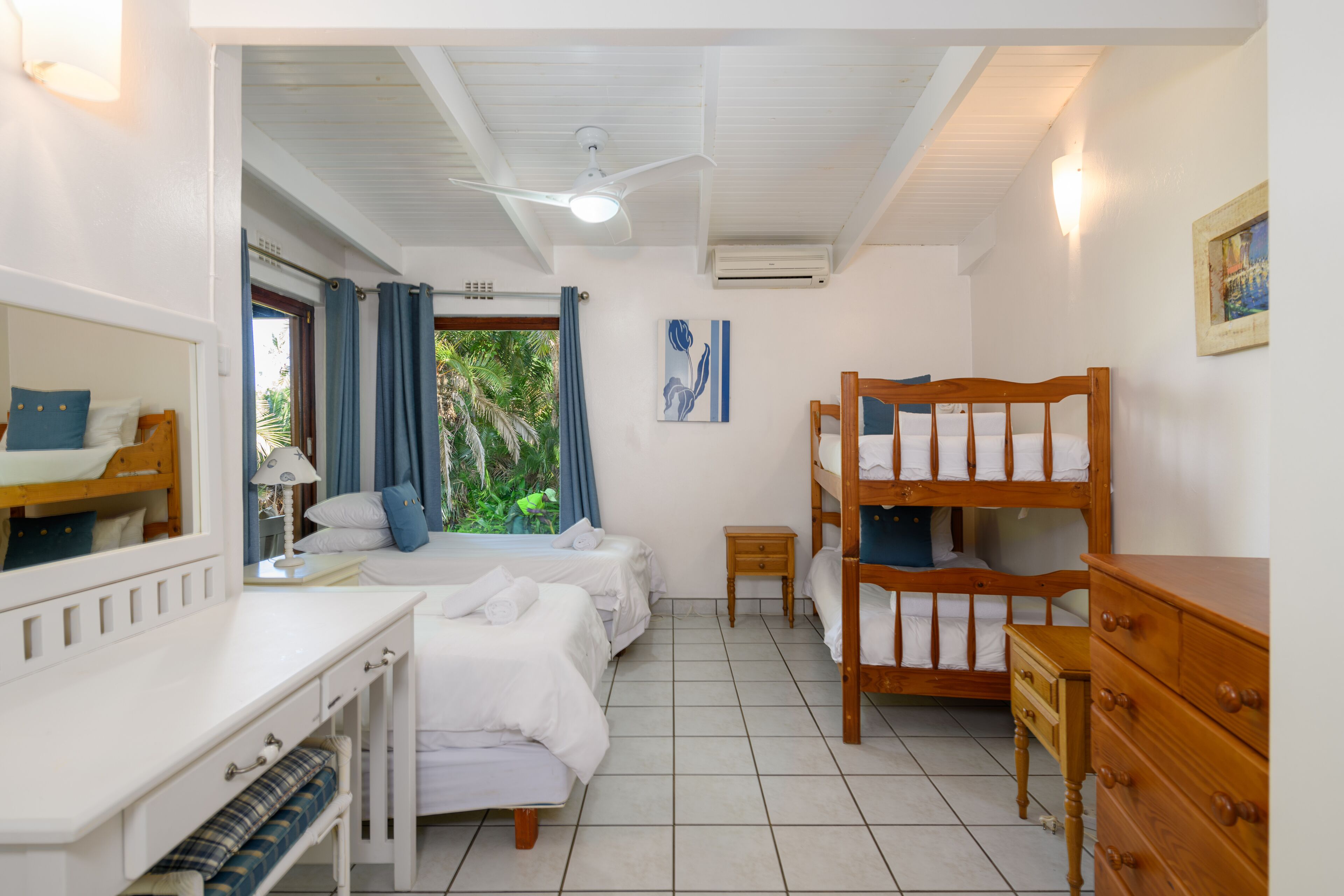 A hotel room with a bunk bed, a desk with a chair, a dresser, a ceiling fan, and a window with curtains.