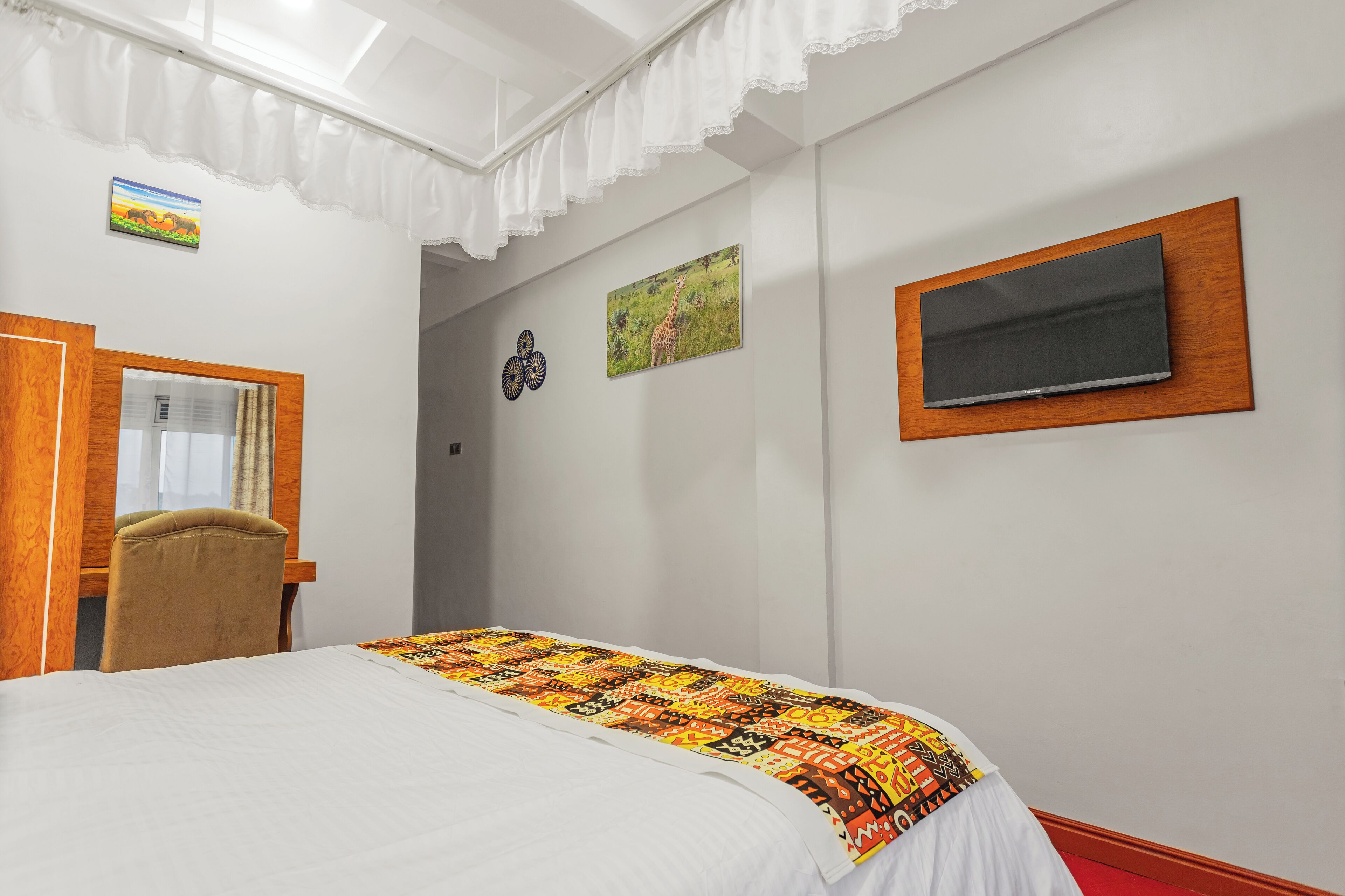 Quarto de hotel com cama, escrivaninha, cadeira, televisão fixada na parede e janela com cortinas.