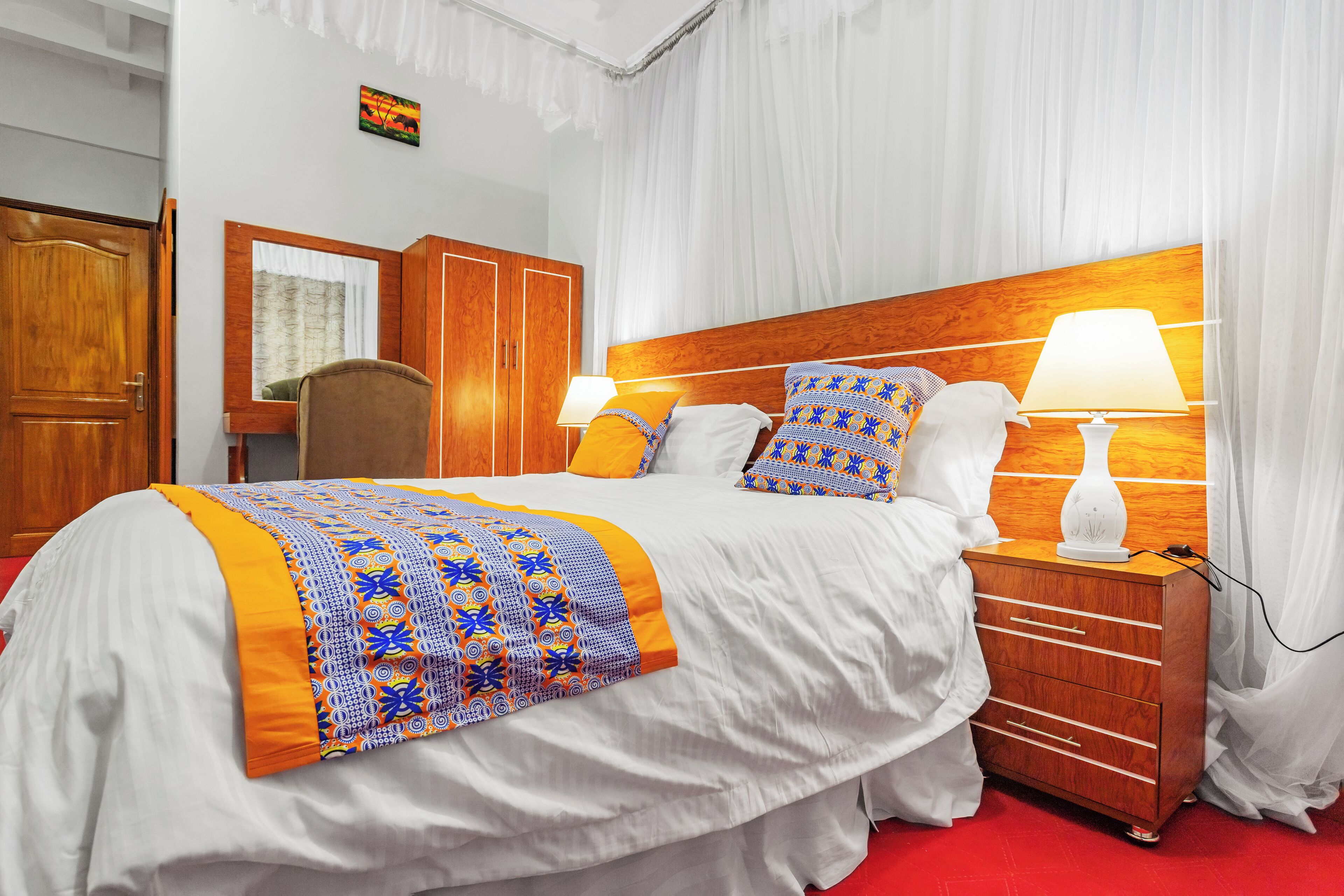 Une chambre d’hôtel avec un grand lit, une tête de lit en bois, des rideaux blancs et une table de chevet avec une lampe.