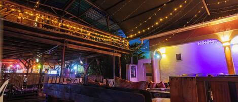 An indoor space with wooden benches and tables, a covered area with string lights, and a wall with a light fixture.