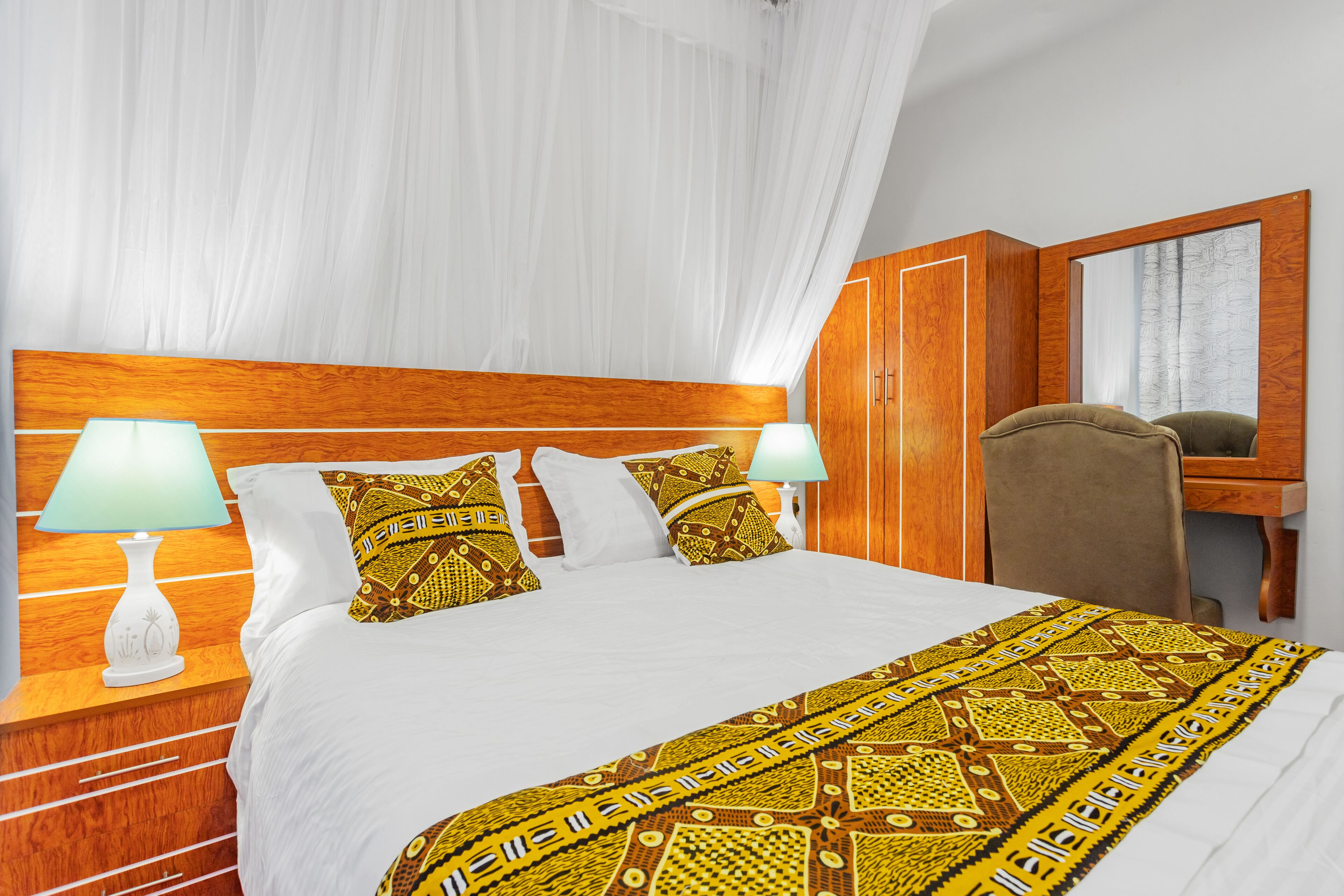 Quarto de hotel com uma cama grande, duas luminárias de cabeceira, um painel de cabeceira de madeira, uma escrivaninha com cadeira e um espelho.