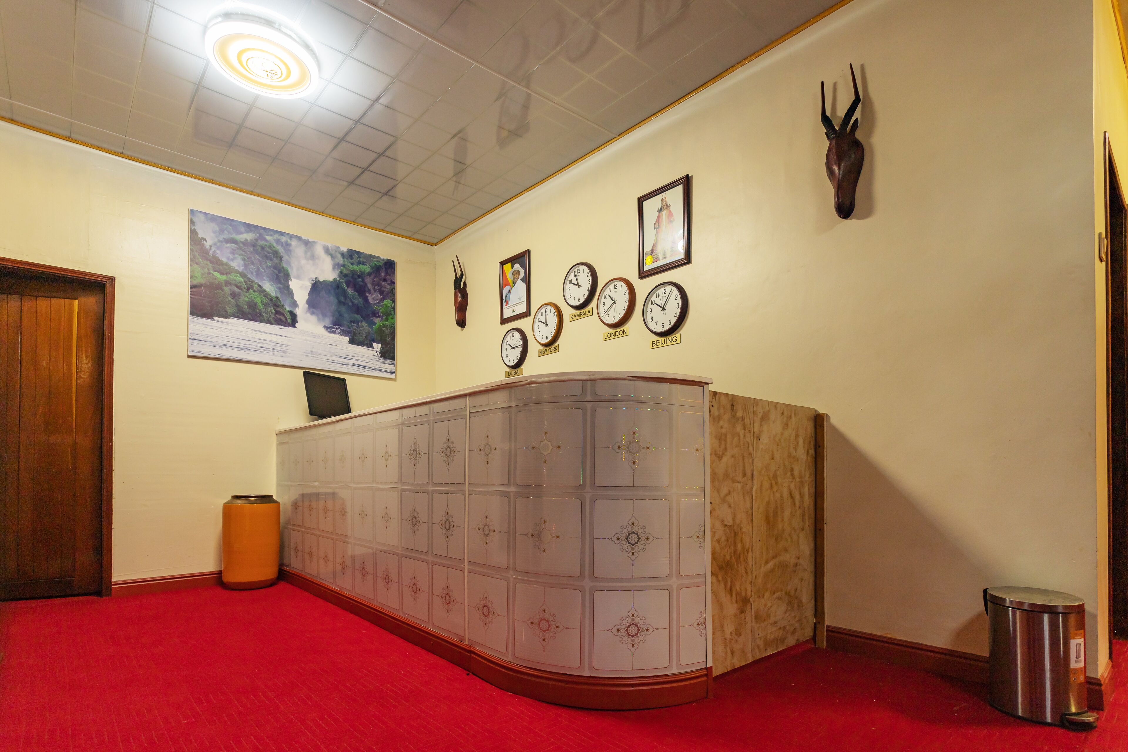 A hotel lobby with a red carpet, a large wooden door, a glass cabinet, wall clocks, a mounted deer head, and a framed picture of a waterfall.
