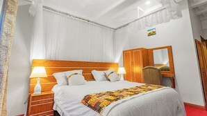Ein ordentlich bezogenes Bett mit weißen Leinen, ein hölzerner Kopfteil und eine farbenfrohe Steudecke. Ein Nachttisch mit einer weißen Lampe und einer kleinen Tischlampe steht daneben. Ein Holzsessel befindet sich neben dem Bett, und ein Spiegel hängt an der Wand.