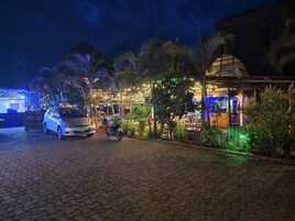 Un camino pavimentado que conduce a un edificio con un área de estar al aire libre cubierta, rodeado de palmeras e iluminación decorativa.