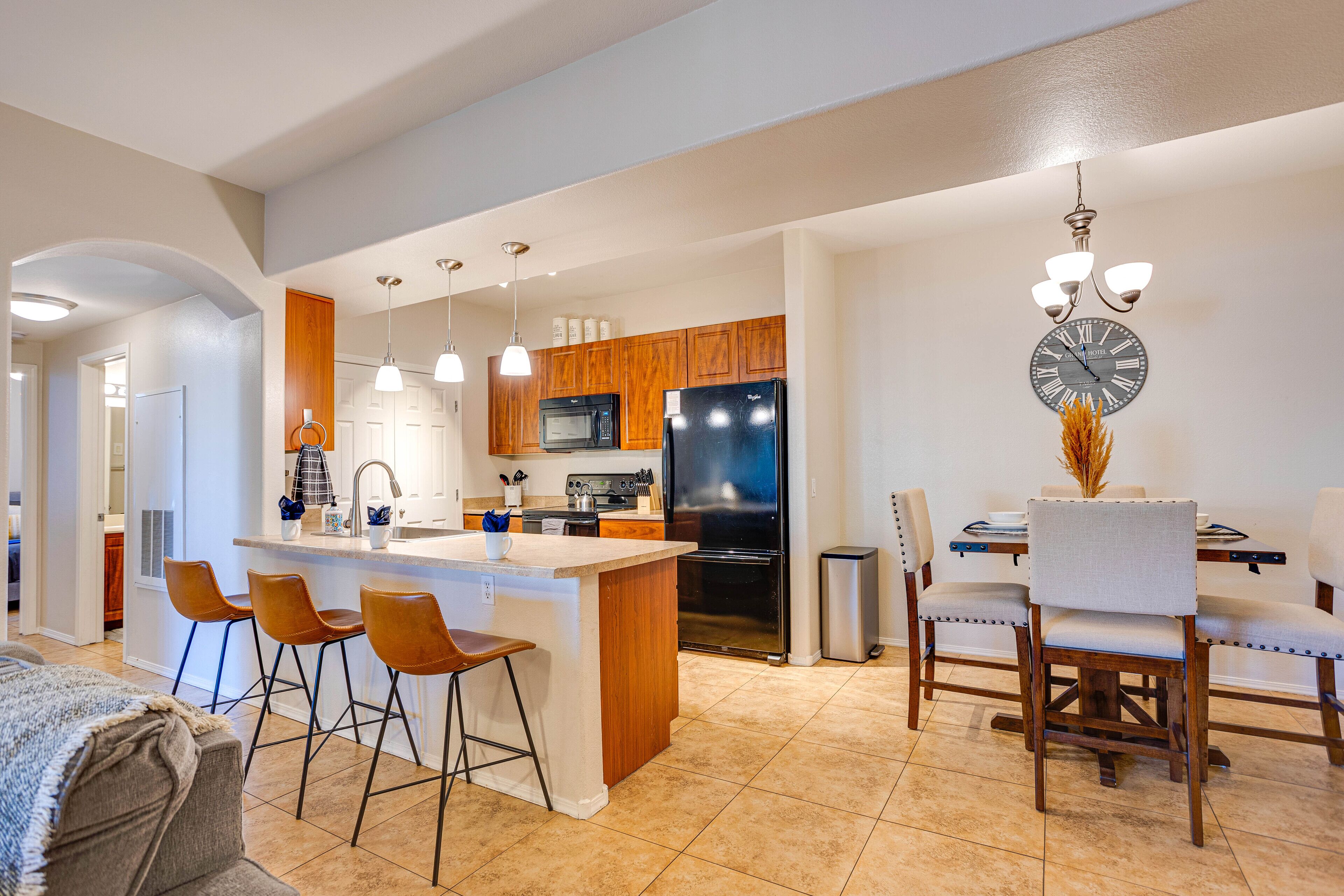 A modern kitchen with a central island, bar stools, wooden cabinets, and a dining area with chairs and a table.