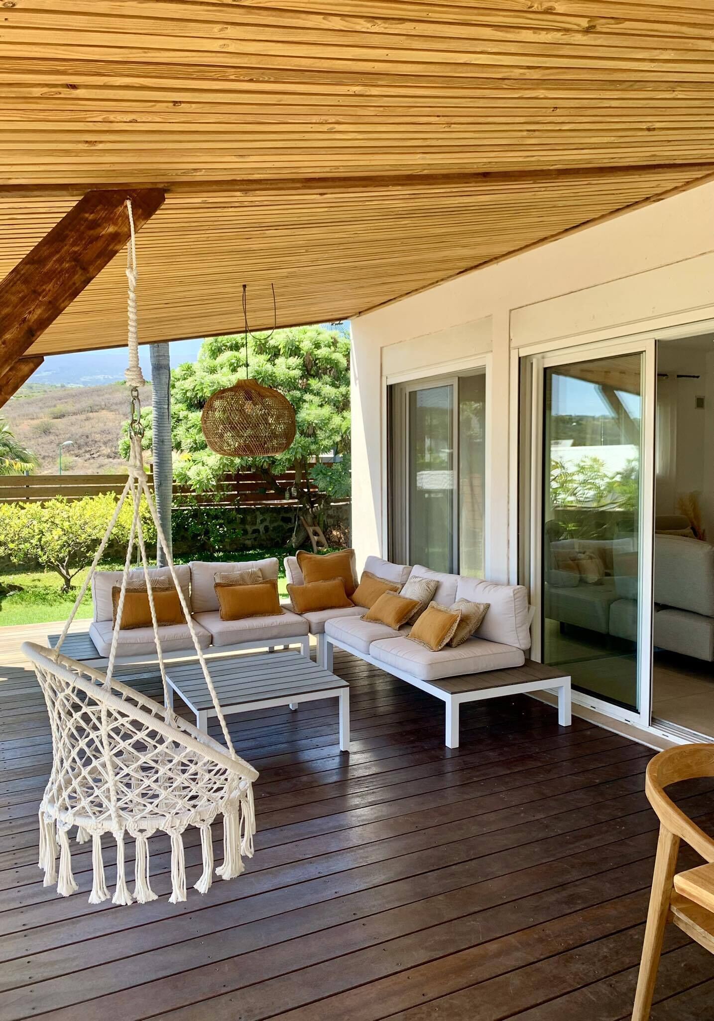 A wooden deck with a white sofa and a hanging chair, large glass doors, and a view of greenery.