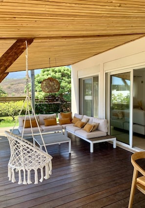 A wooden deck with a white sofa and a hanging chair, large glass doors, and a view of greenery.