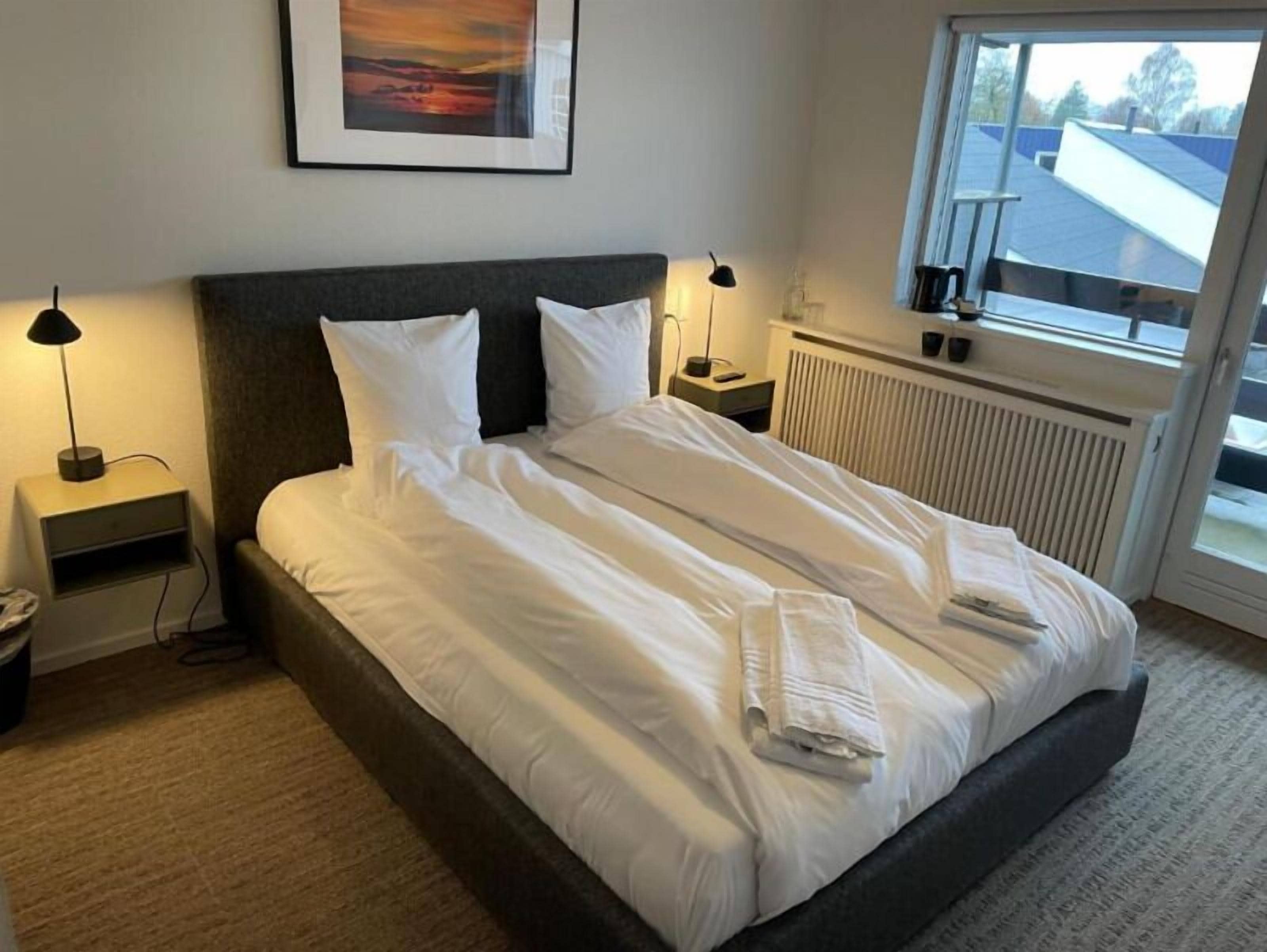 A neatly made bed with white linens and a folded towel, a nightstand with a lamp, a framed picture on the wall, and a window with a view of buildings.