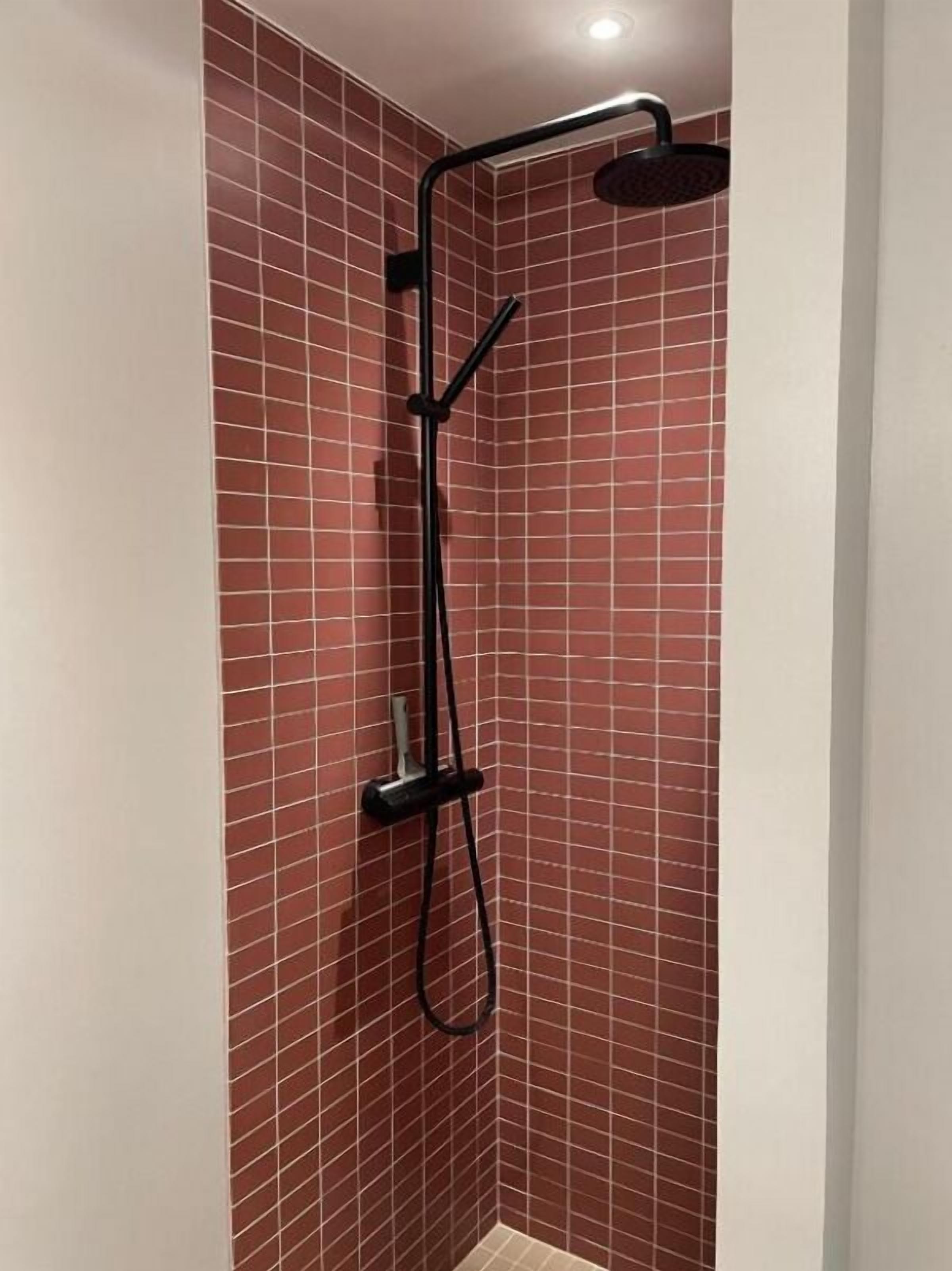 A shower with a black hose and a handheld showerhead, set against a red-tiled wall.