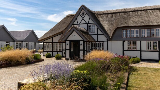 A traditional half-timbered house with a thatched roof, a sign reading "KRO", and a paved courtyard with various plants and shrubs. - Hotel Nørrevang (Vaeggerlose)