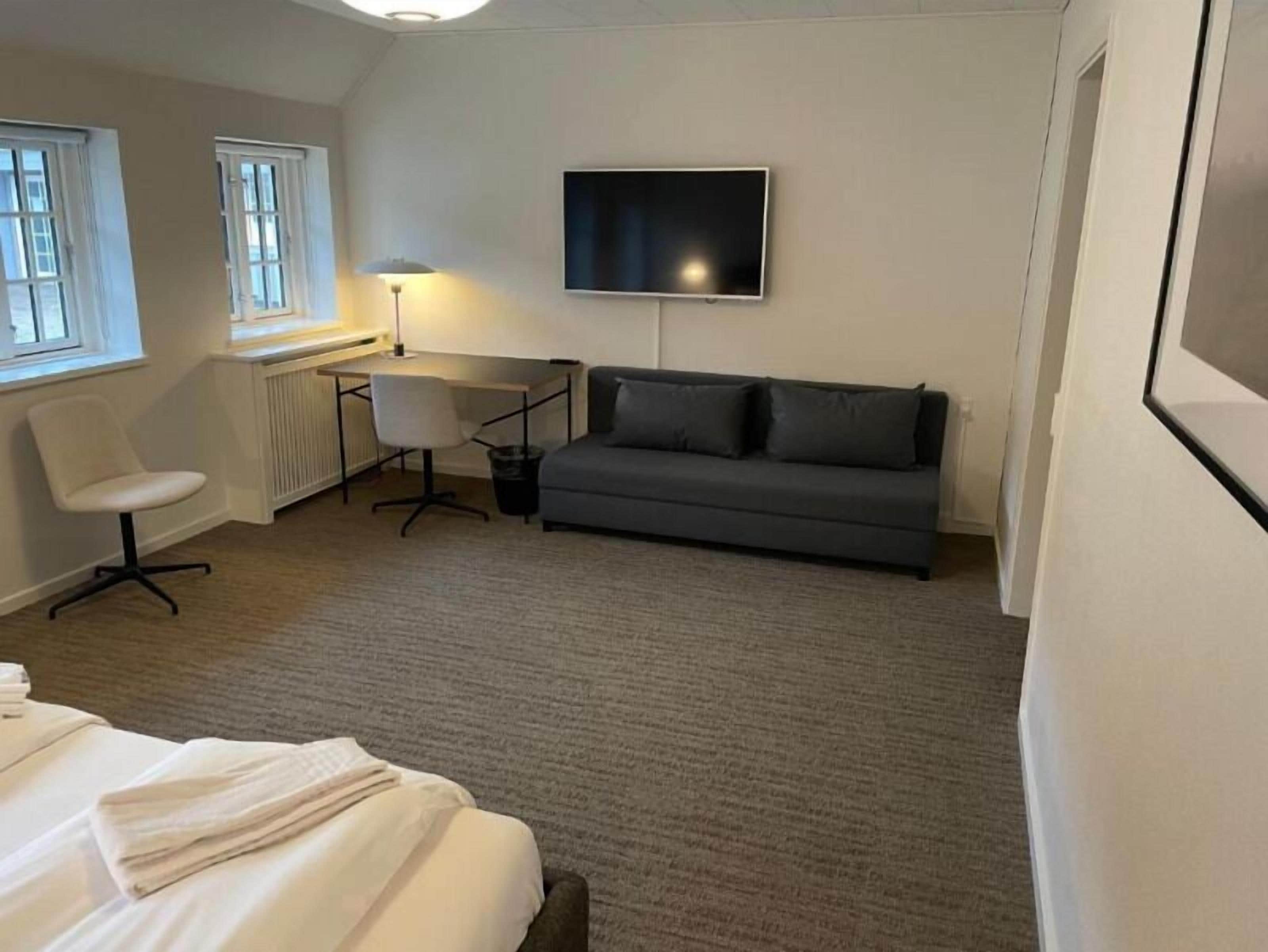 A hotel room with a bed, a grey sofa, a desk with a chair, a TV, and a window.