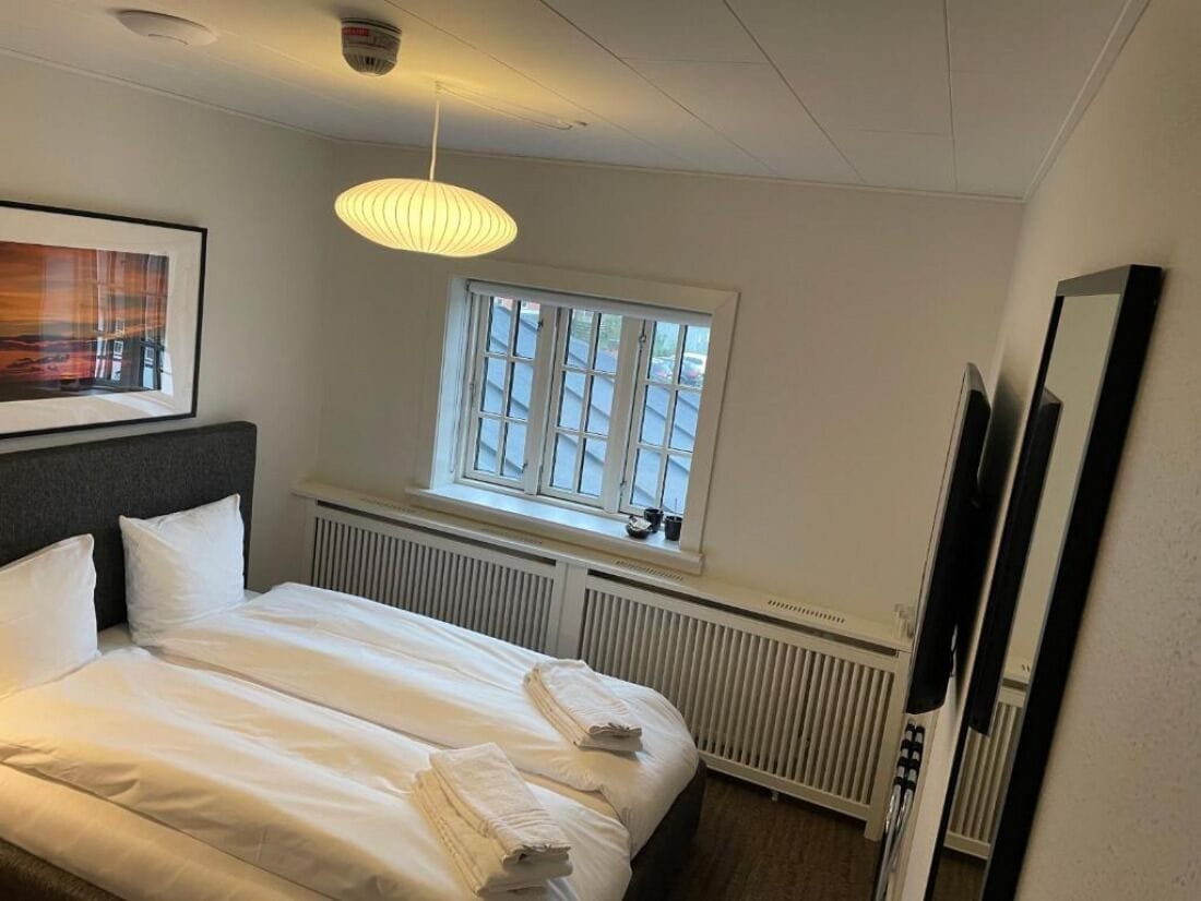 A neatly made bed with white linens and towels, a window with blinds, a radiator, and a framed picture on the wall.