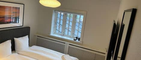 Una cama bien hecha con sábanas y toallas blancas, una ventana con persianas, un radiador y un cuadro en la pared.