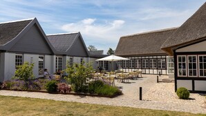 A courtyard with outdoor seating, a thatched-roof building, and a white building with large windows. - Hotel Nørrevang (Vaeggerlose)