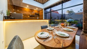 Un espace repas moderne avec une table en bois dressée pour quatre personnes, un comptoir de cuisine agrémenté d’une plante et une vue sur l’extérieur grâce à de grandes fenêtres.