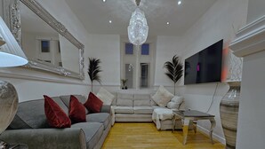 A living room with a grey sofa, white armchair, and a small table. There is a large mirror on the wall, a chandelier, and a flat-screen TV mounted on the wall. - White Tulip Central 3bedroom Duplex Loft (Liverpool)