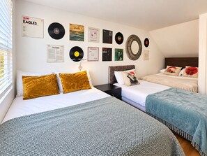 A hotel room with two beds, a wall decorated with vinyl records and posters, and a window with blinds.