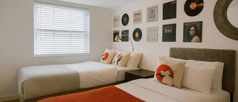 A hotel room with two beds, a large window with blinds, wall-mounted records, and a round mirror.