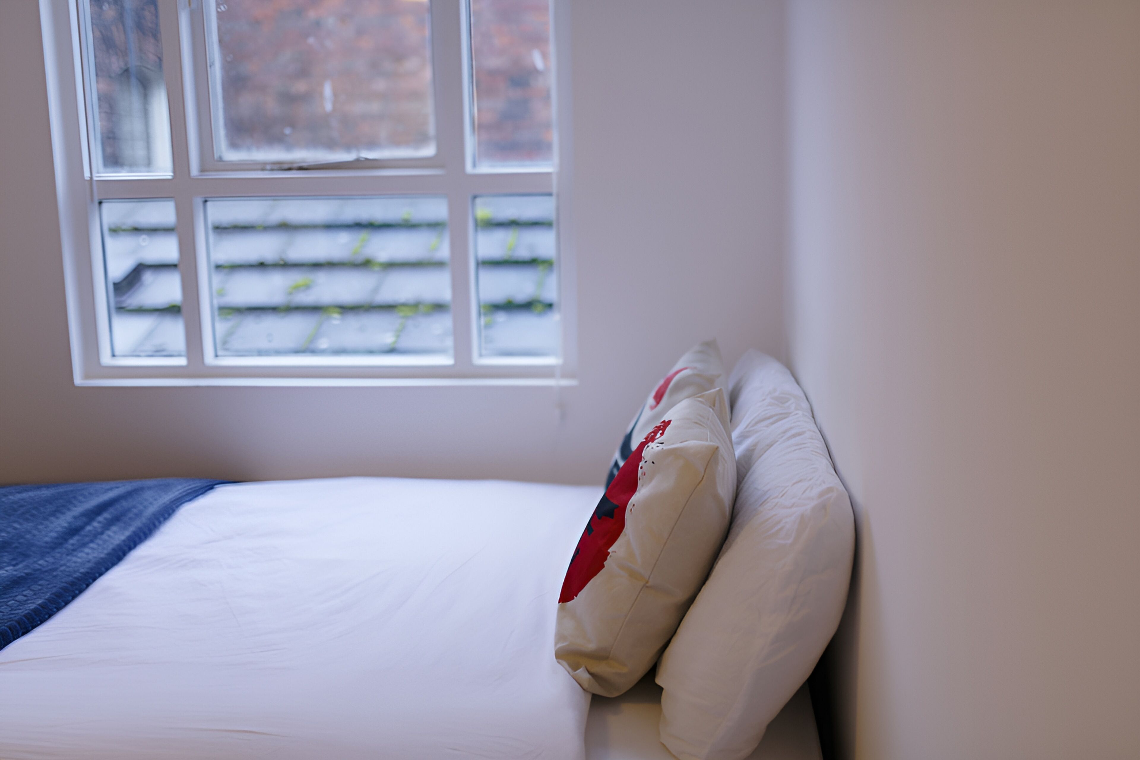 Ein Bett mit weißer Bettwäsche und einer blauen Tagesdecke, ein Fenster mit Blick auf eine Backsteinmauer und ein weißes Kopfkissen mit rotem Muster.