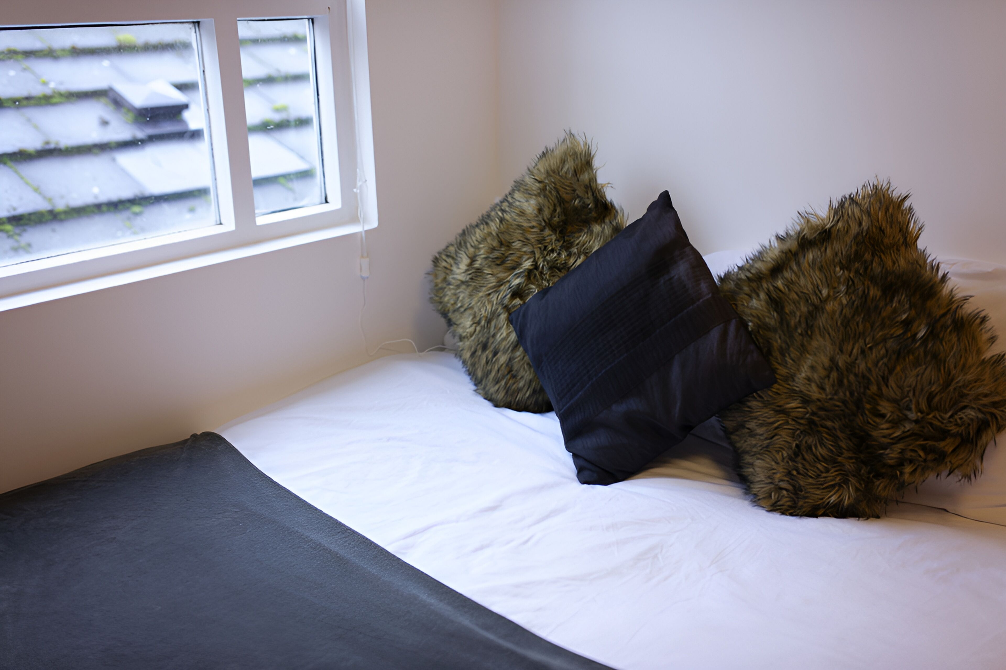 Cama com lençóis brancos e duas almofadas fofas, uma escura e outra marrom clara, em um quarto com janela.