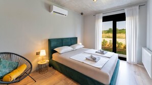 A neatly made bed with white linens and towels, a green upholstered headboard, a small round table with a lamp, and a wicker chair with a cushion. - Stunning home in Pula (Pula)