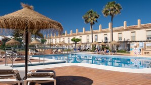 Zona de piscina con sillones de descanso, una sombrilla de paja, palmeras y un edificio con balcones.