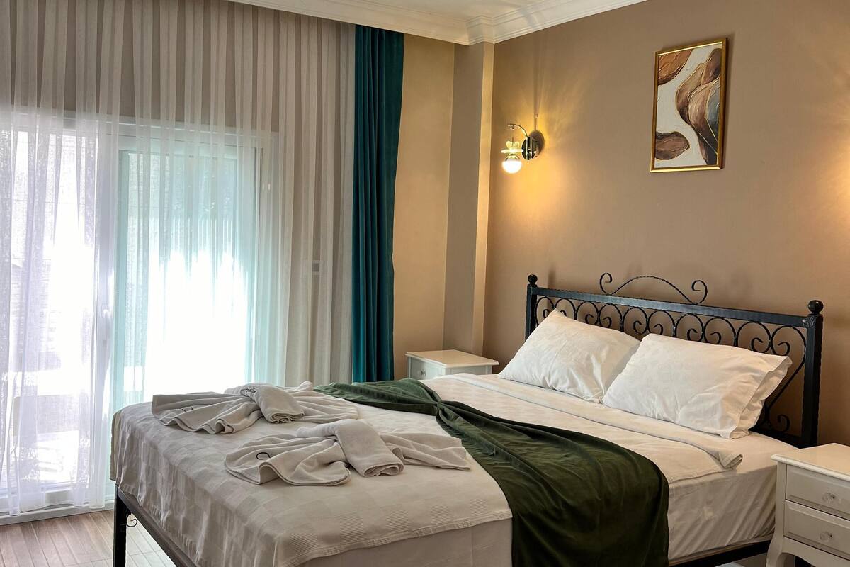 A hotel room with a bed, bedside tables, a painting, and a window with curtains.