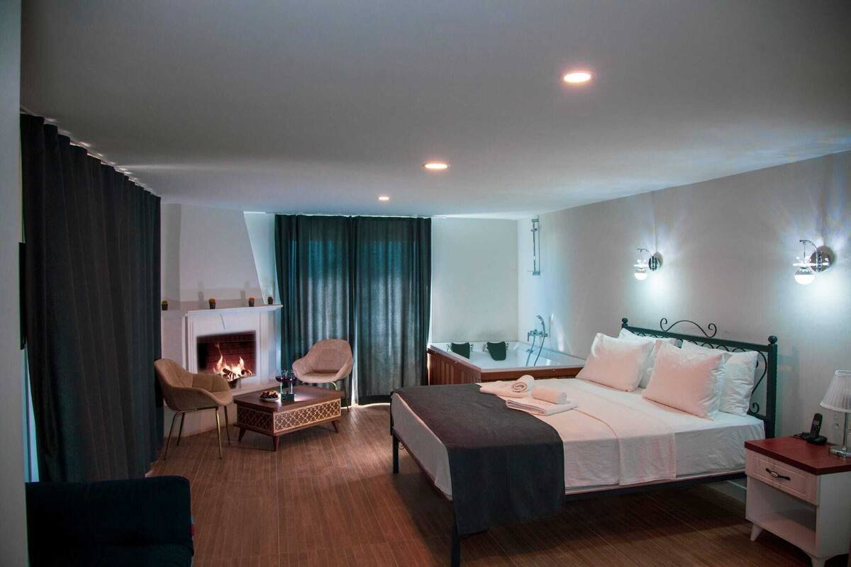 A hotel room with a bed, a sitting area with a chair and a small table, a fireplace, a bathroom with a bathtub, and a nightstand with a lamp.