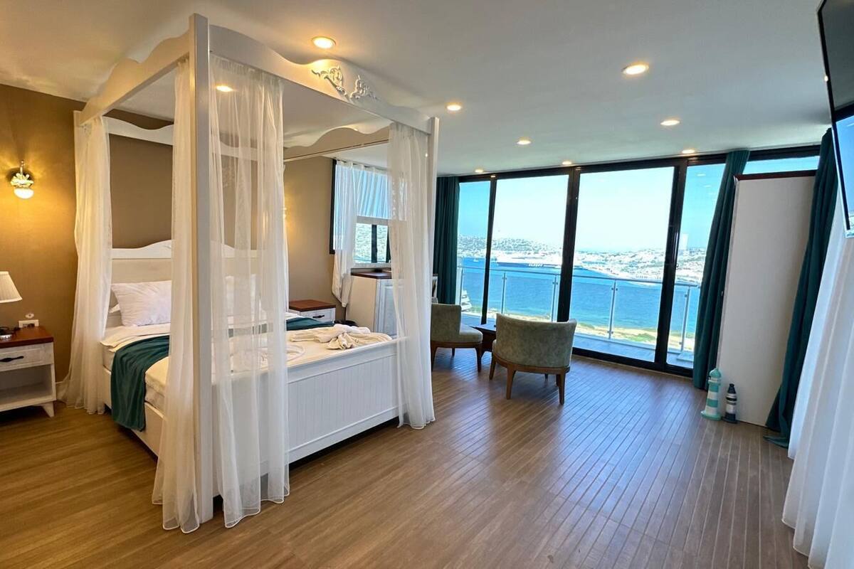 A bedroom with a four-poster bed, a nightstand, a chair, and a view of the sea.