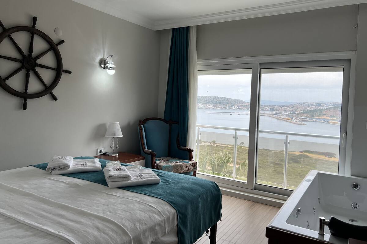 A hotel room with a bed, a large window with a view of a body of water and a city, a ship's wheel on the wall, and a bathtub.