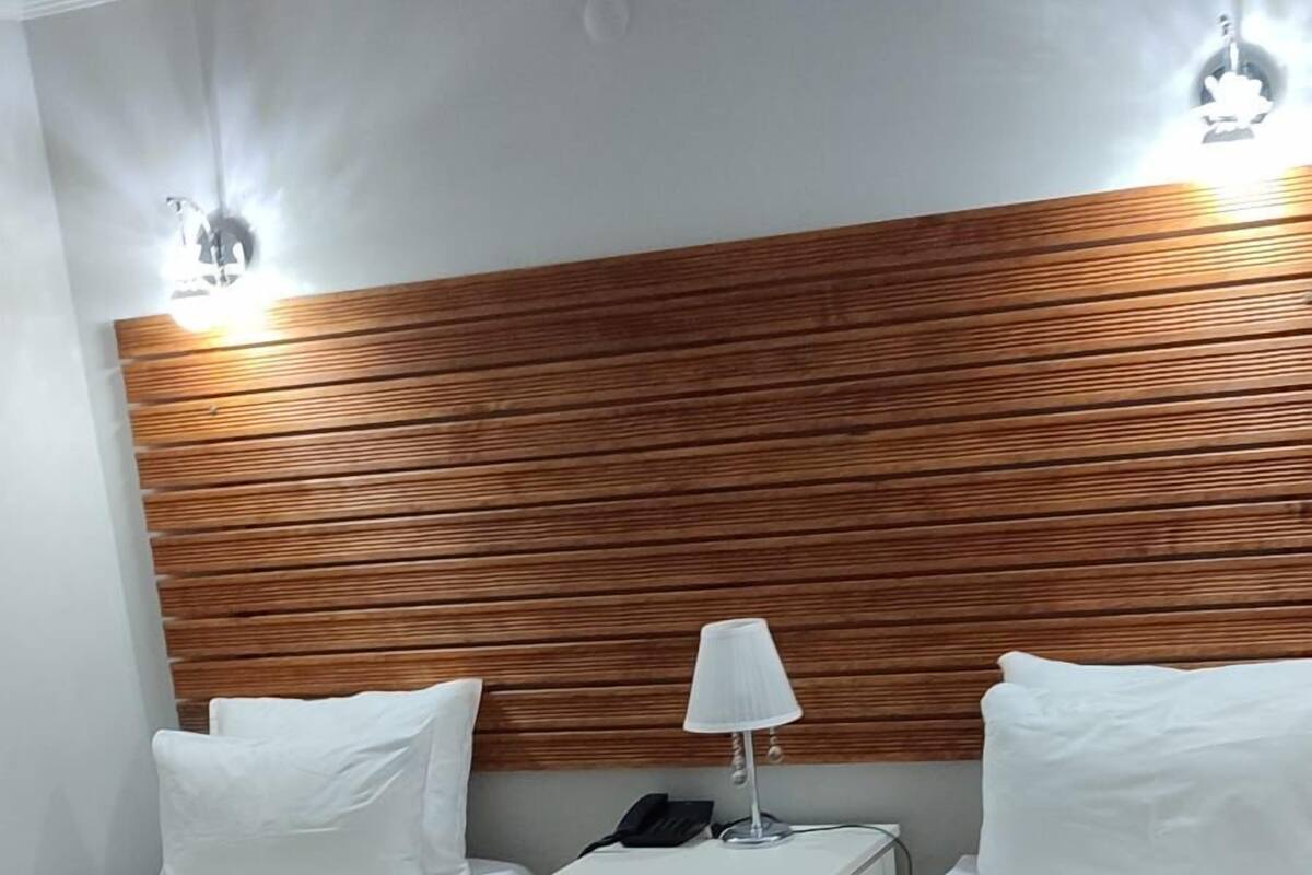 A hotel room with two beds, a wooden headboard, a bedside table with a lamp, and a green throw blanket.