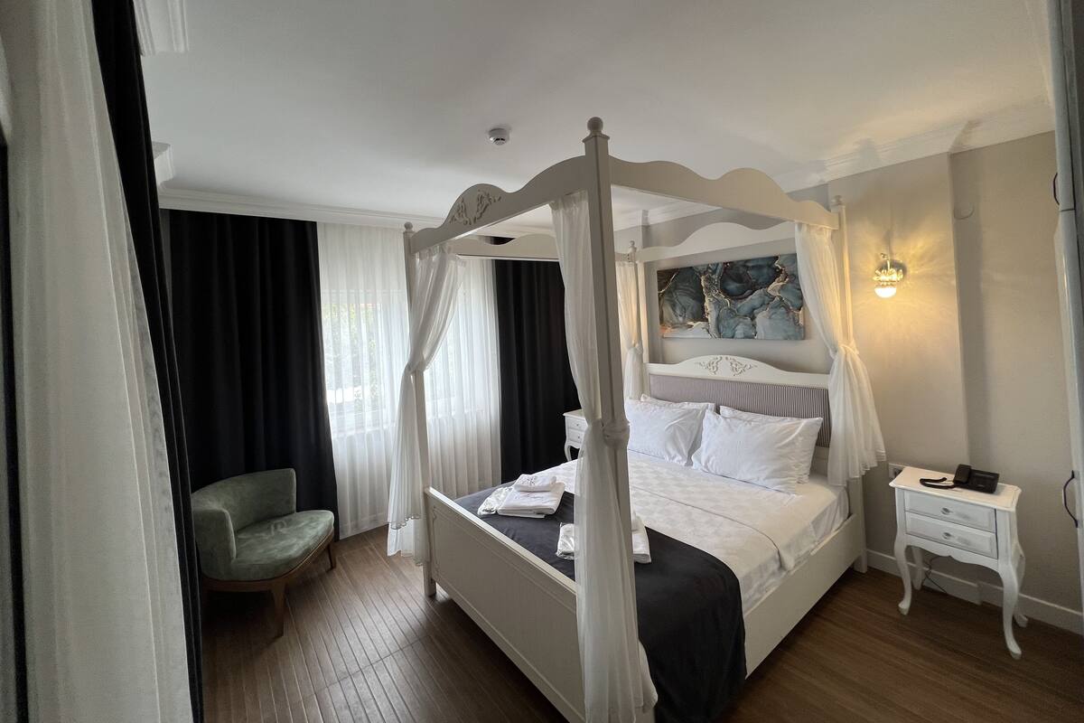 A four-poster canopy bed with white bedding, a bedside table with a lamp, and a chair in a hotel room.