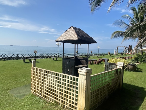 A gazebo with a thatched roof, surrounded by a wooden fence, overlooking a grassy area with benches and a view of the sea.