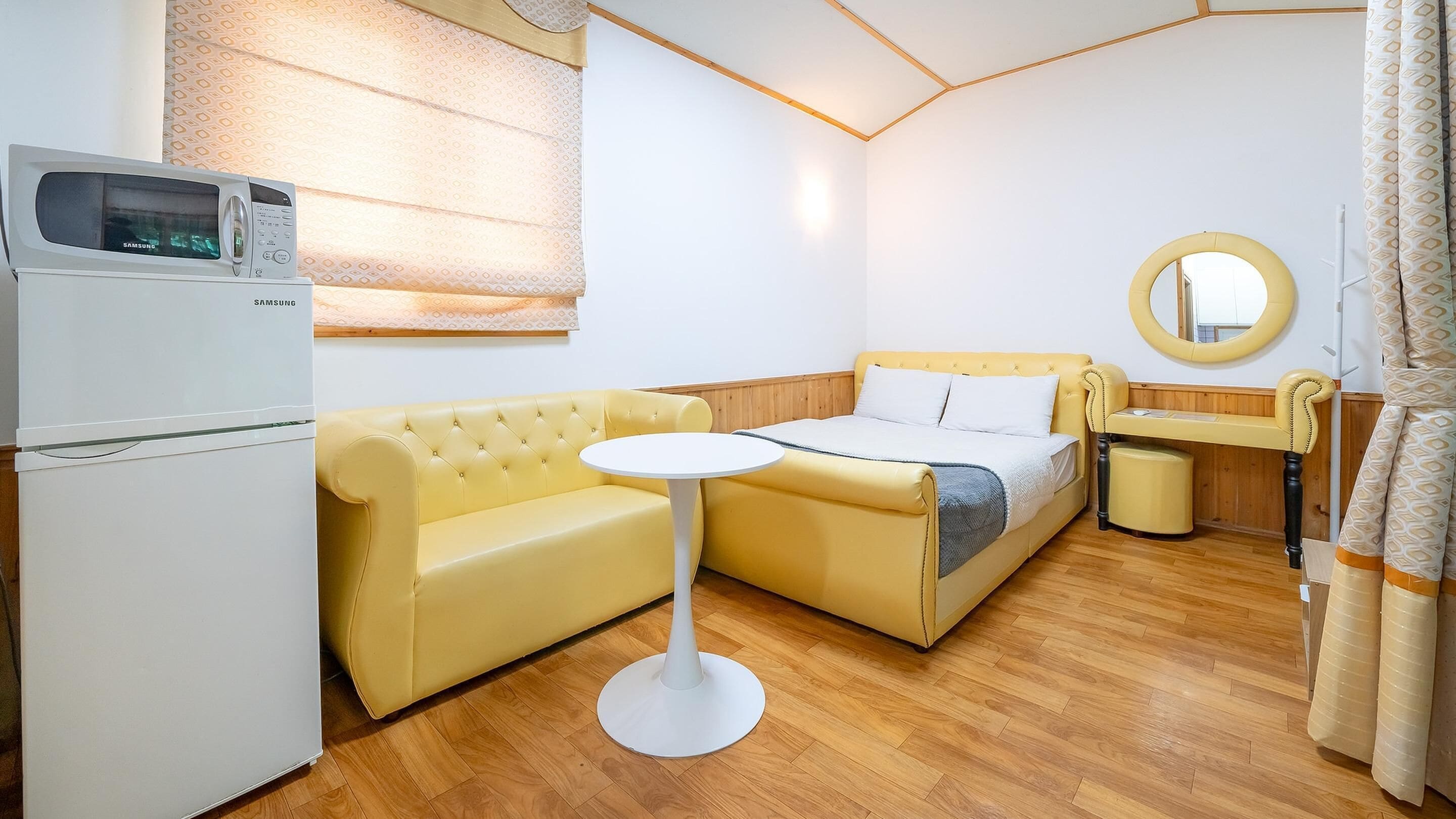 A compact room with a yellow sofa, a small round table, a bed, a microwave, and a mirror.
