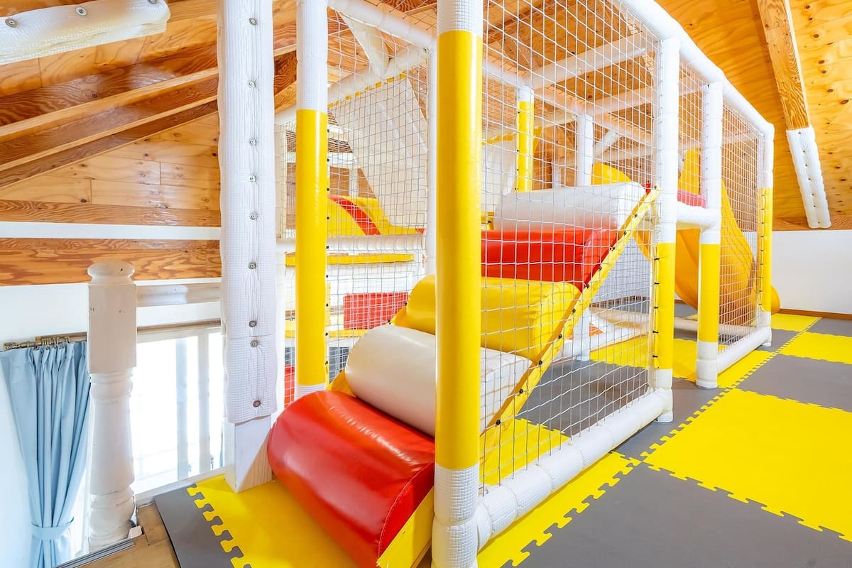 A playpen with yellow and red padding, a wooden ceiling, and a window with blue curtains.