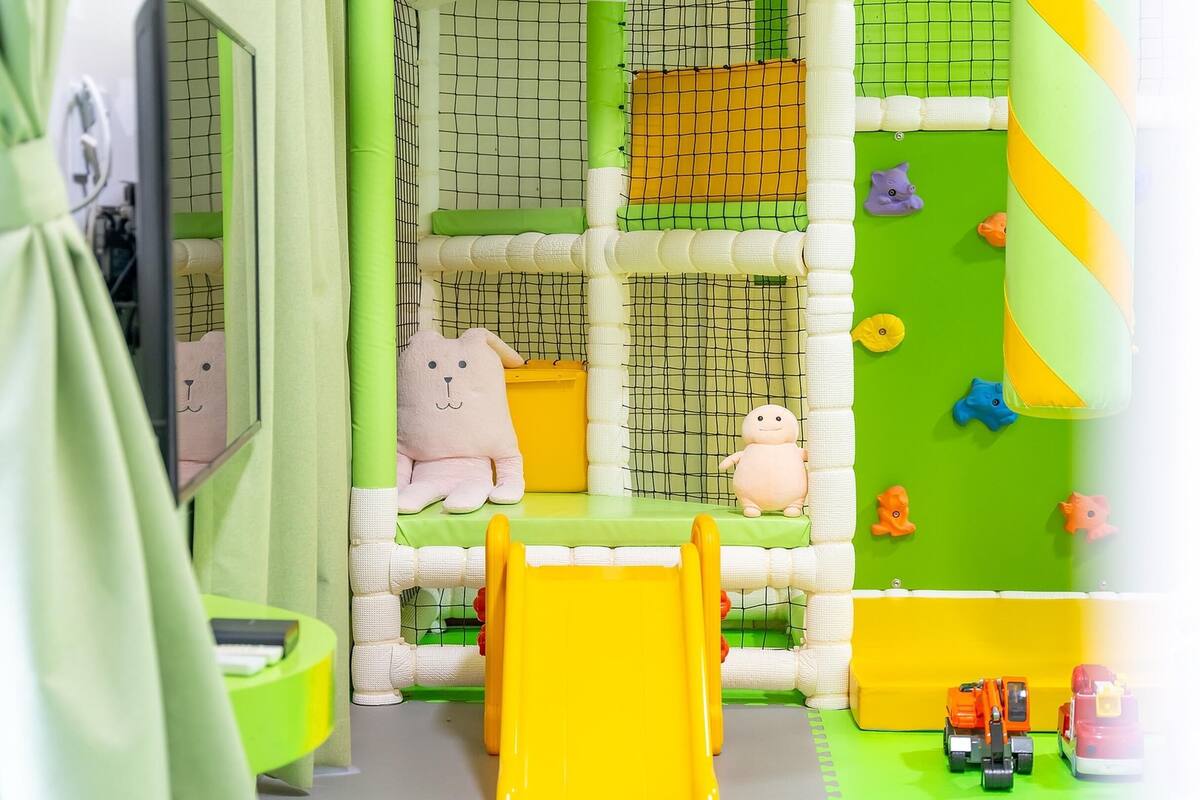 A children's play area with a netted climbing wall, soft play structures, and a yellow slide.