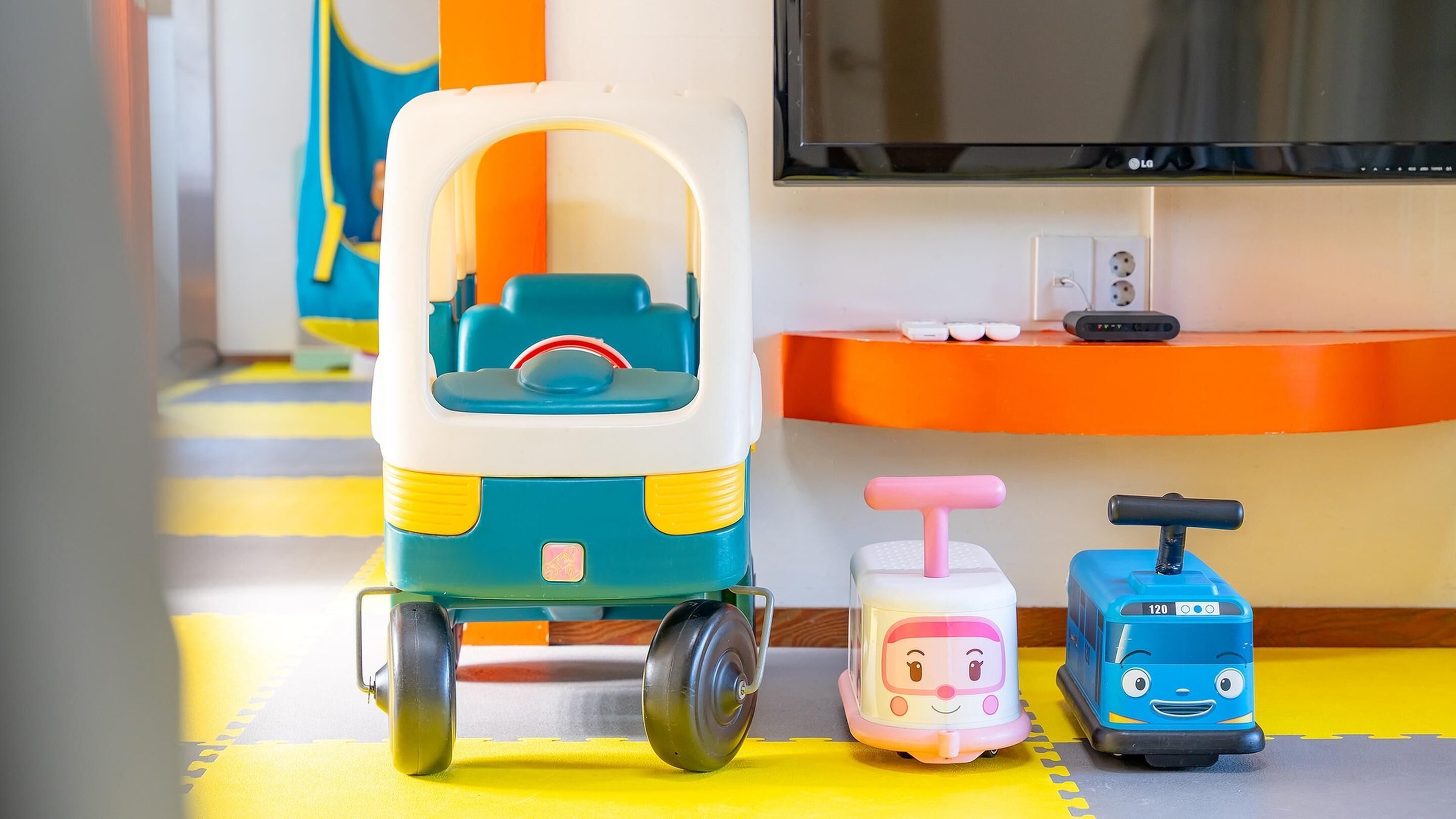 A toy train set on a yellow mat with a TV and orange shelf in the background.