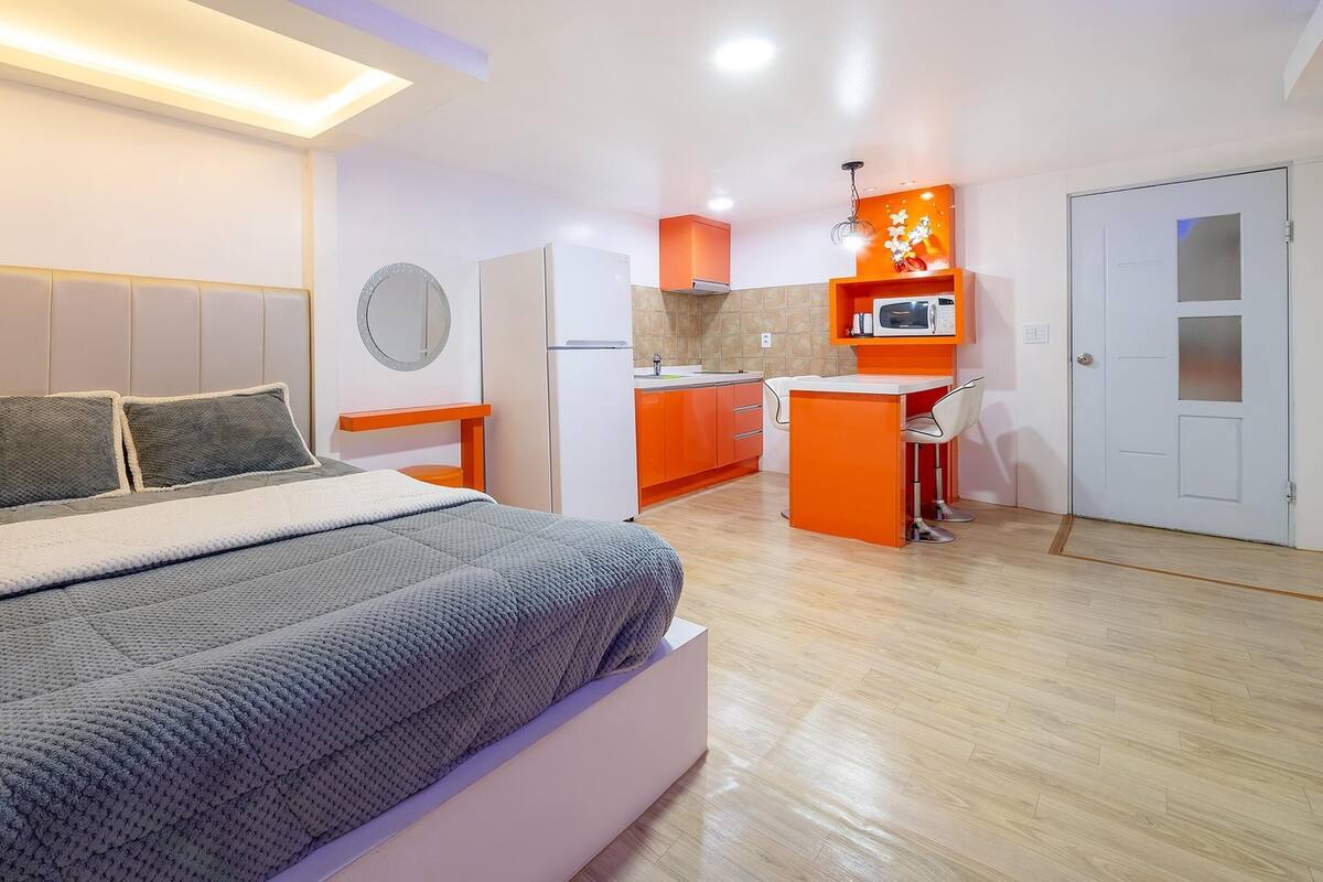 A modern bedroom with a bed, a bedside table, and a small kitchen area with a microwave and orange cabinets.