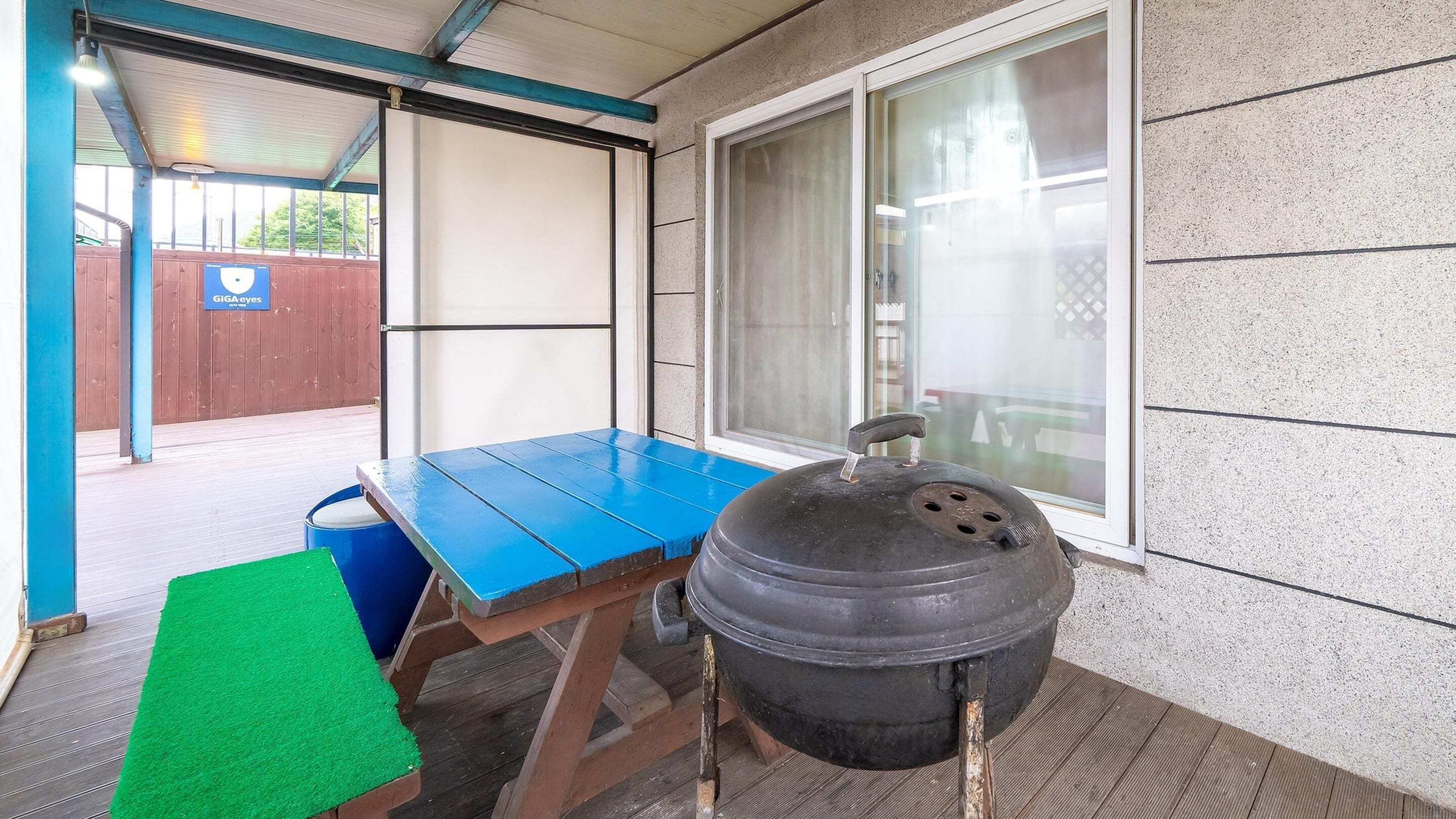 A covered patio area with a blue table, a barbecue grill, and a green mat.
