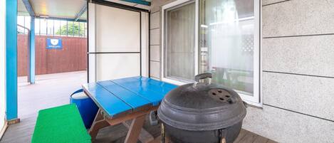 A covered patio area with a blue table, a barbecue grill, and a green mat.