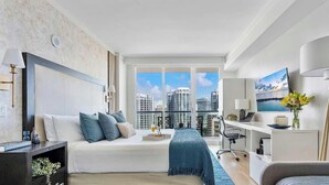 A modern bedroom with a bed, desk, and large windows offering a city view. - Coastal Art Studio Suite with Workspace (Miami)