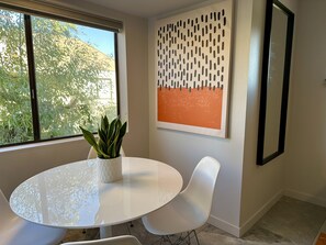 Een moderne kamer met een ronde witte tafel, twee witte stoelen, een potplant, een kunstwerk aan de muur en een raam met uitzicht op bomen.