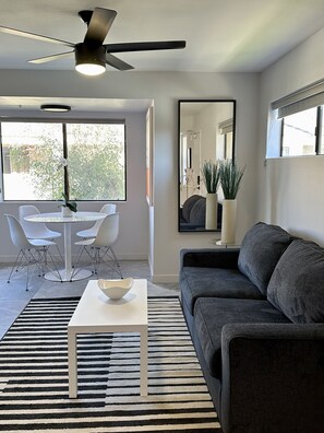 A modern living room with a gray sofa, a white coffee table, a dining area with a white table and chairs, a ceiling fan, and a large window with a view of greenery. - Chic Condo fully remodeled  in the heart of Palm Springs.  (Palm Springs)