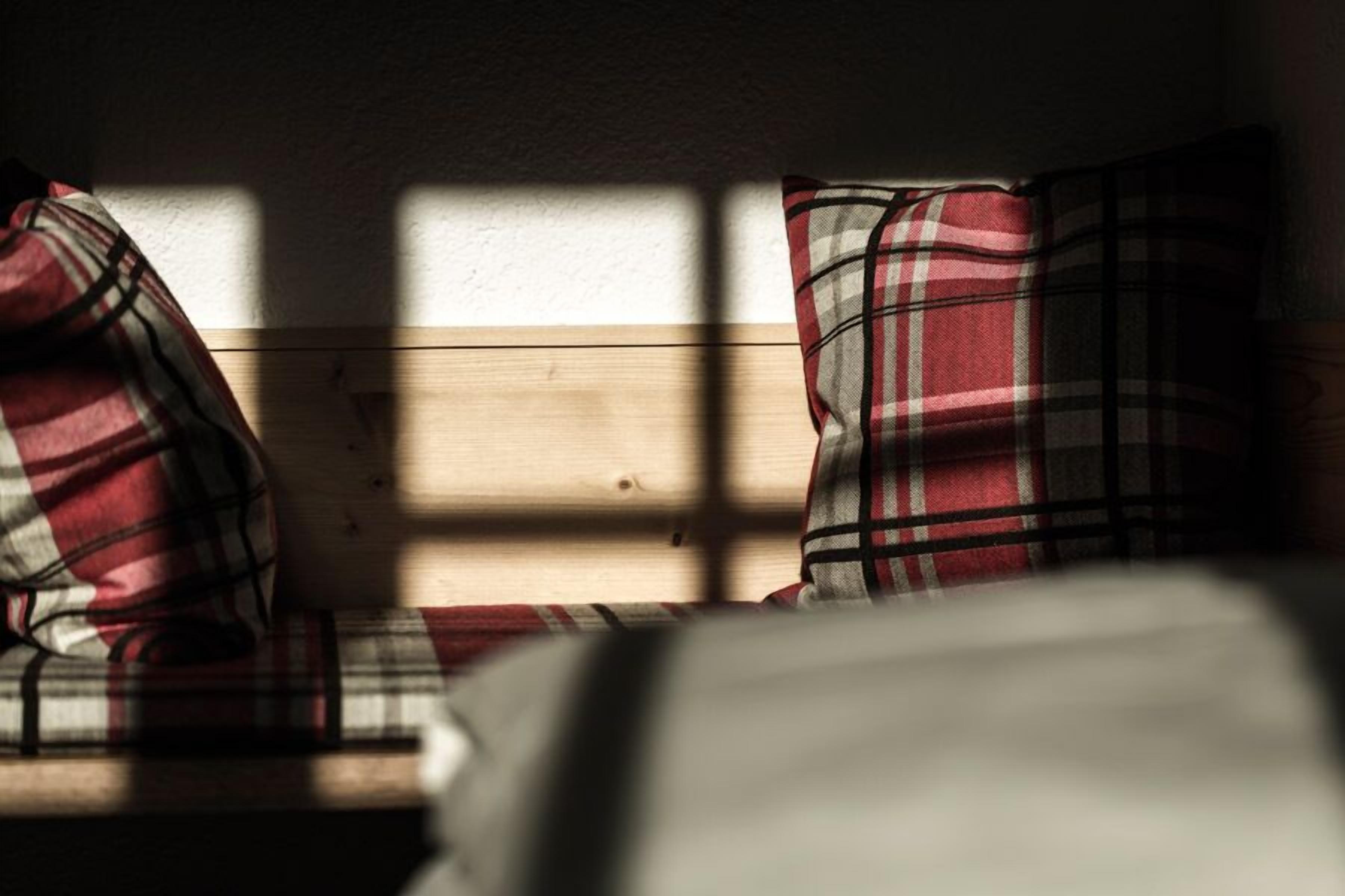 Un banco de madera con cojines a cuadros en una habitación con una pared lisa y una ventana que proyecta sombras.