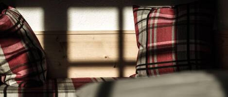 A wooden bench with plaid cushions in a room with a plain wall and a window casting shadows.