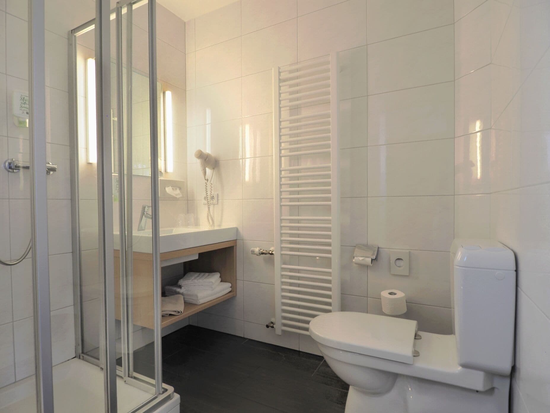 Un baño moderno con cabina de ducha de vidrio, inodoro blanco, toallero y repisa para toallas.